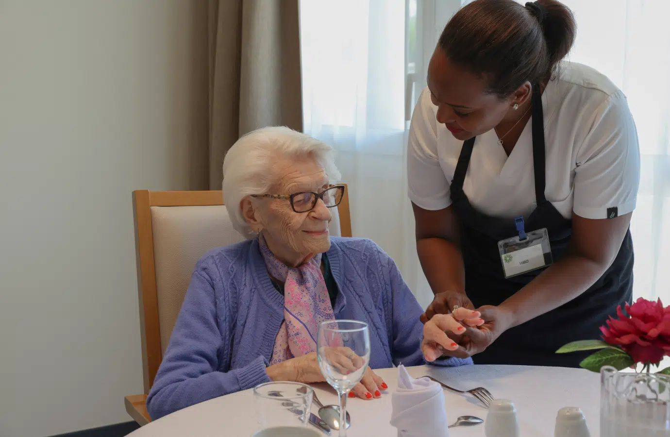 Femme âgée assise à une table est assistée par une soignante | Clinalliance voisins-le-bretonneux
